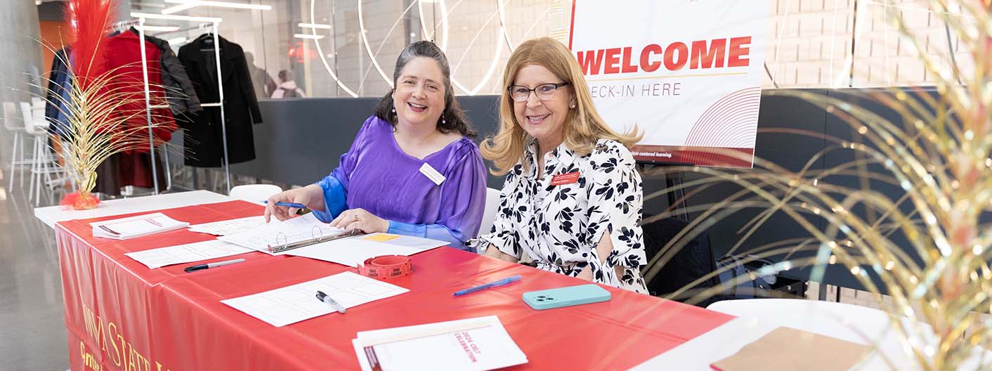 The image shows two women sitting at a welcome desk, smiling at the camera. The desk is covered with a red tablecloth that has "Iowa State University" printed on it. There are several papers, pens, and a phone on the desk. Behind them, a large sign reads "WELCOME - CHECK-IN HERE." The setting appears to be a modern indoor space with some decorations and clothing racks in the background.