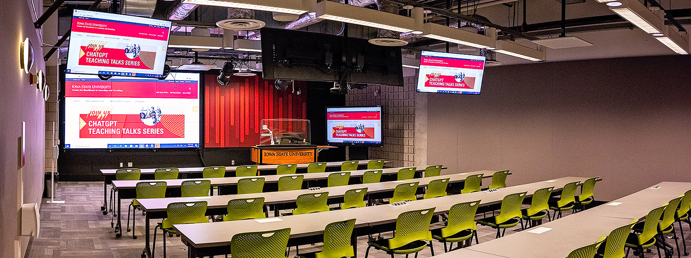 The image shows a modern lecture or presentation room at Iowa State University. The room features rows of tables and green chairs arranged in a classroom setup. At the front of the room is a podium with the Iowa State University logo, and multiple large screens are displaying a presentation titled "ChatGPT Teaching Talks Series." The walls and ceiling are equipped with advanced lighting and audio-visual equipment, indicating a high-tech environment designed for interactive and multimedia presentations.