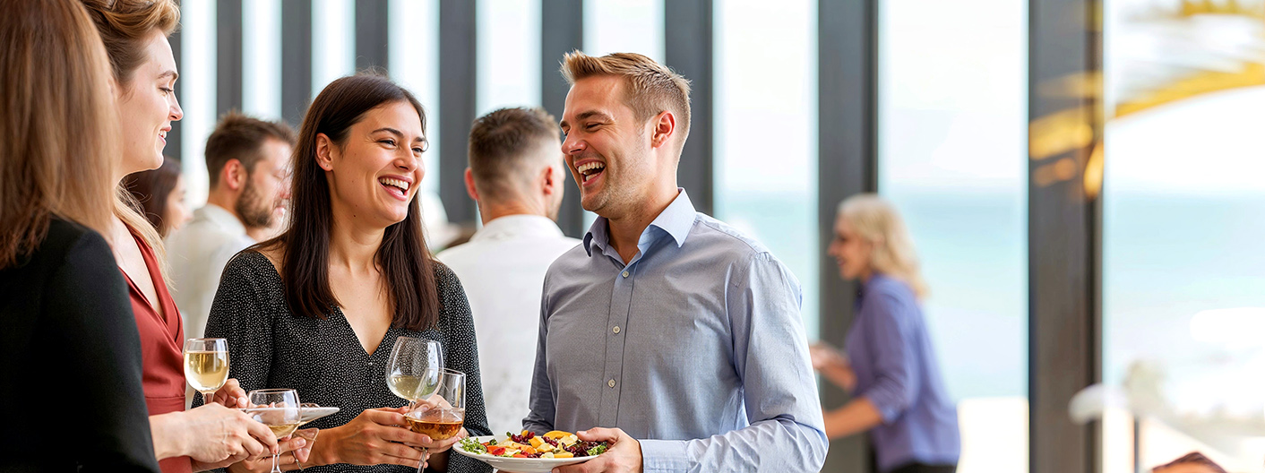 A group of three people, two women and one man, are standing together, smiling and chatting while holding drinks and plates of food at a social event. They are dressed in business-casual attire, and the background features other people mingling in a bright, modern setting with large windows.