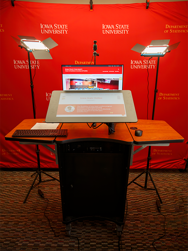 The image shows a professional recording setup at Iowa State University, Department of Statistics. It features a podium with a large touchscreen monitor displaying the university's website, a keyboard, a mouse, and two lighting panels positioned on either side for optimal illumination. A red backdrop with "Iowa State University Department of Statistics" branding is visible behind the setup.